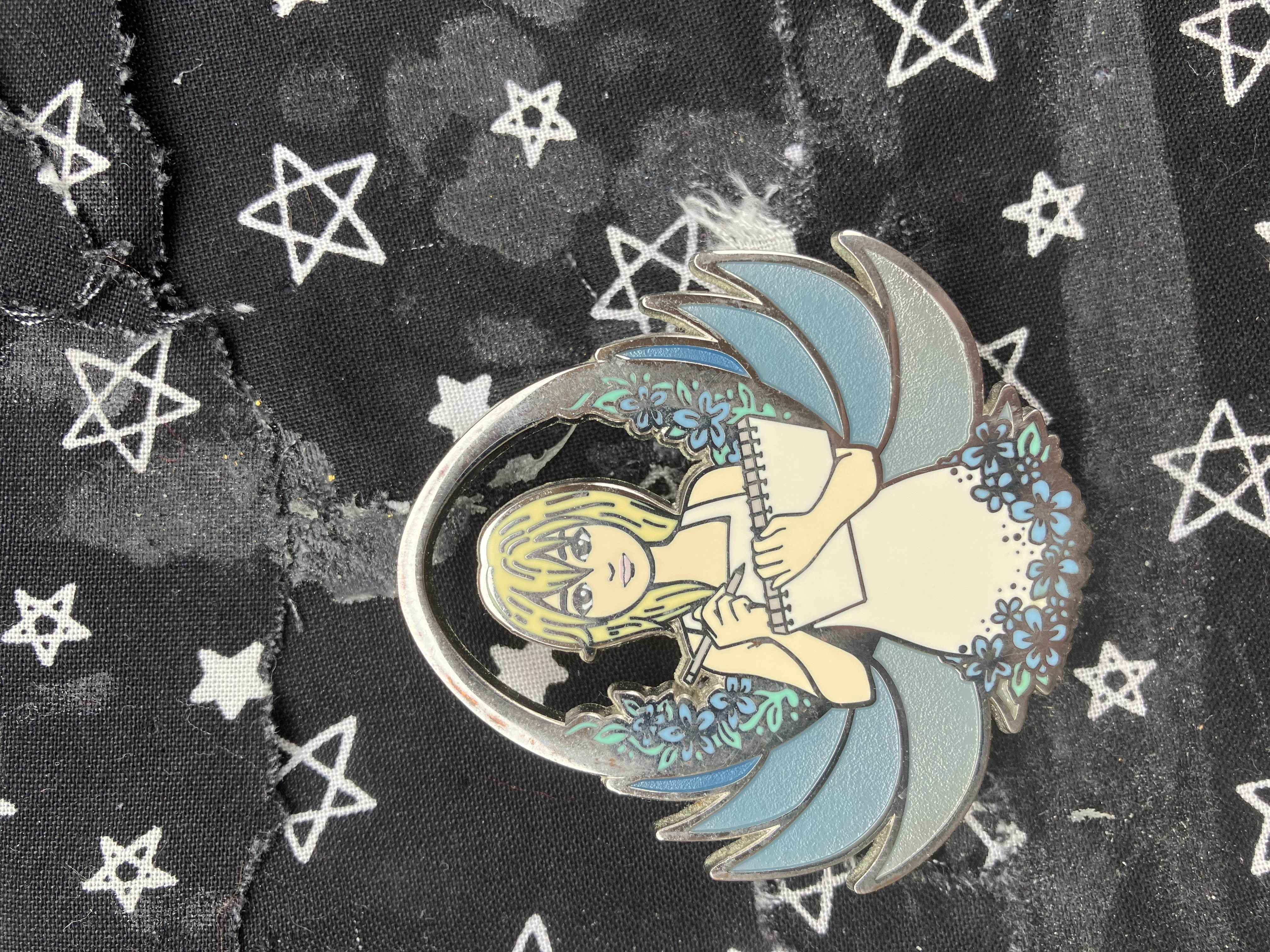 Enamel pin of Kingdom hearts Namine holding a sketchpad and a color pencil in her hand with a open pod and blue flowers behind her.