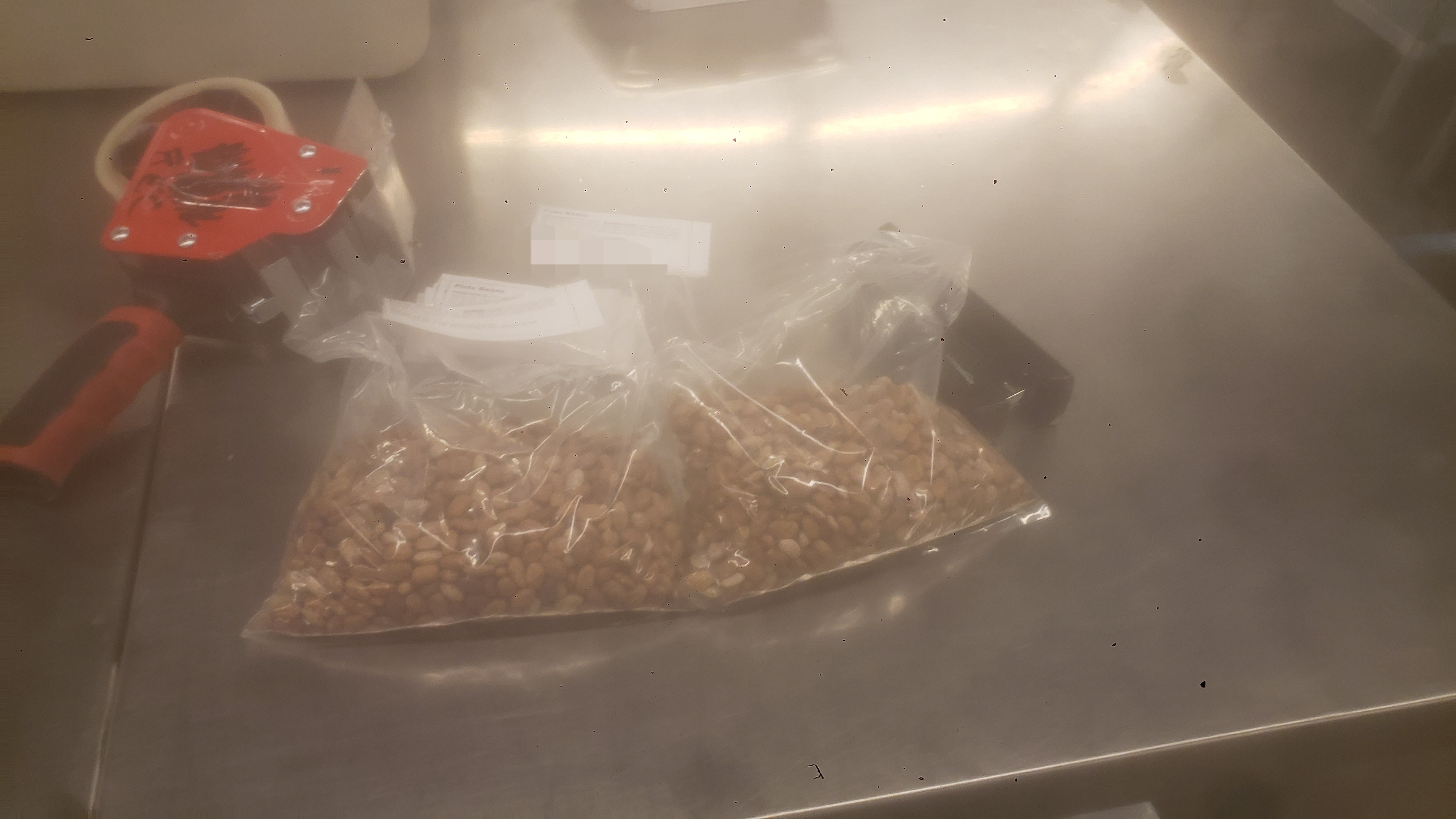 One bag of pinto bags with a label and another without one next to it with labels and a red tape gun sitting on top of it.