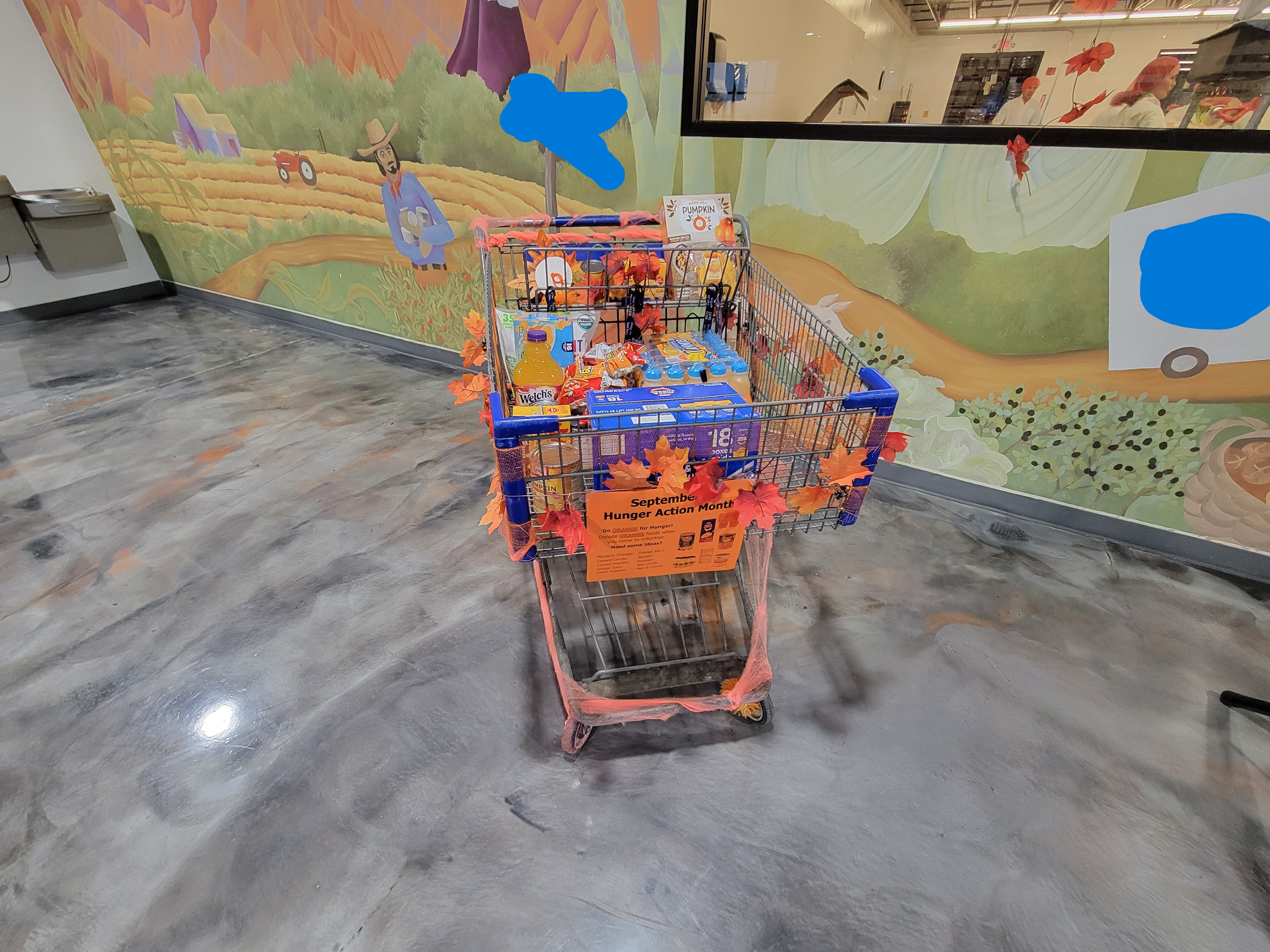Big shopping cart decorated with autumn decorations full of orange-ish foods and has a sign talking donating orange foods for hungry action month.