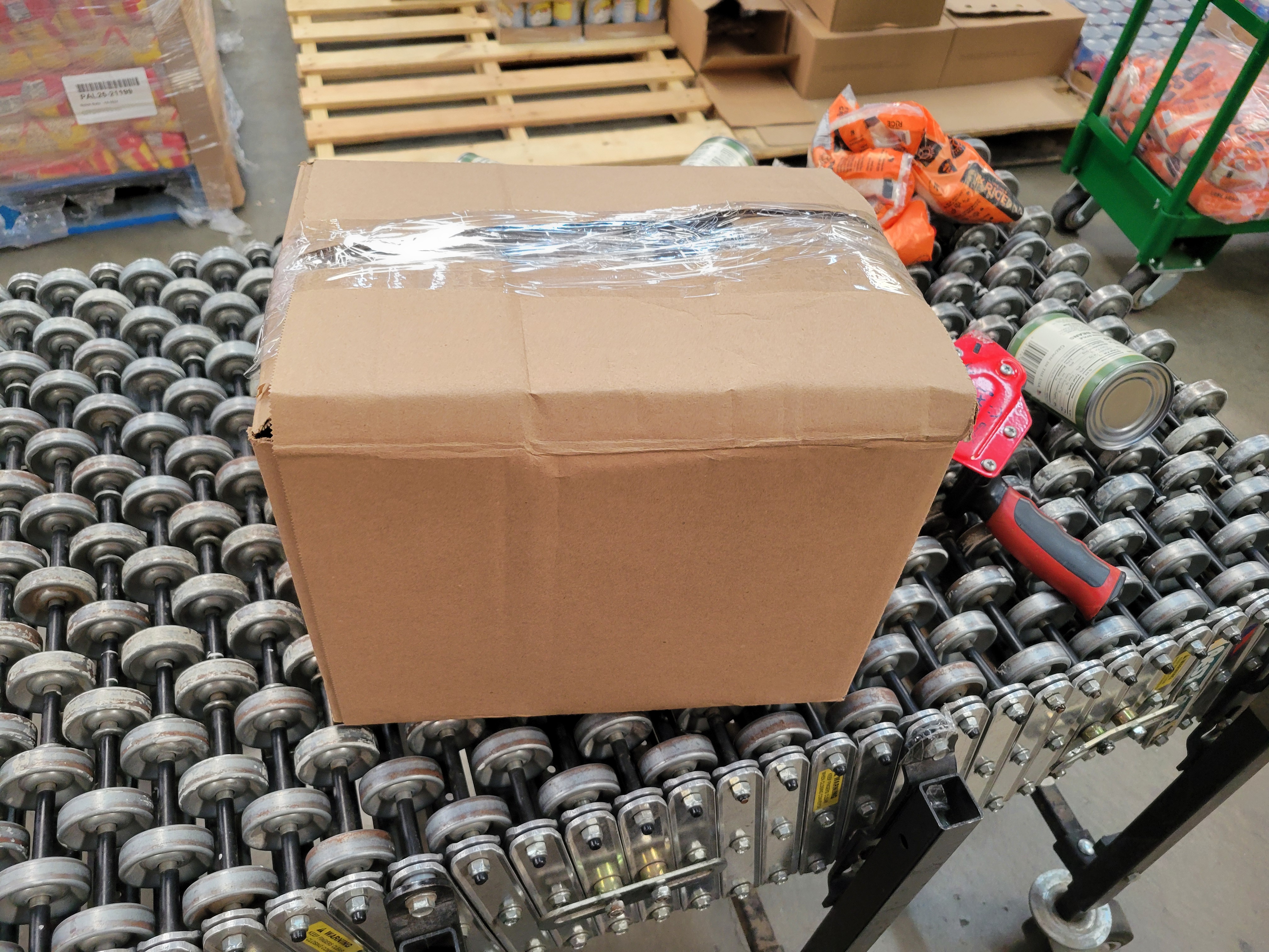 Taped box on conveyor belt next to a tape gun, canned green beans and bags of rice behind it.