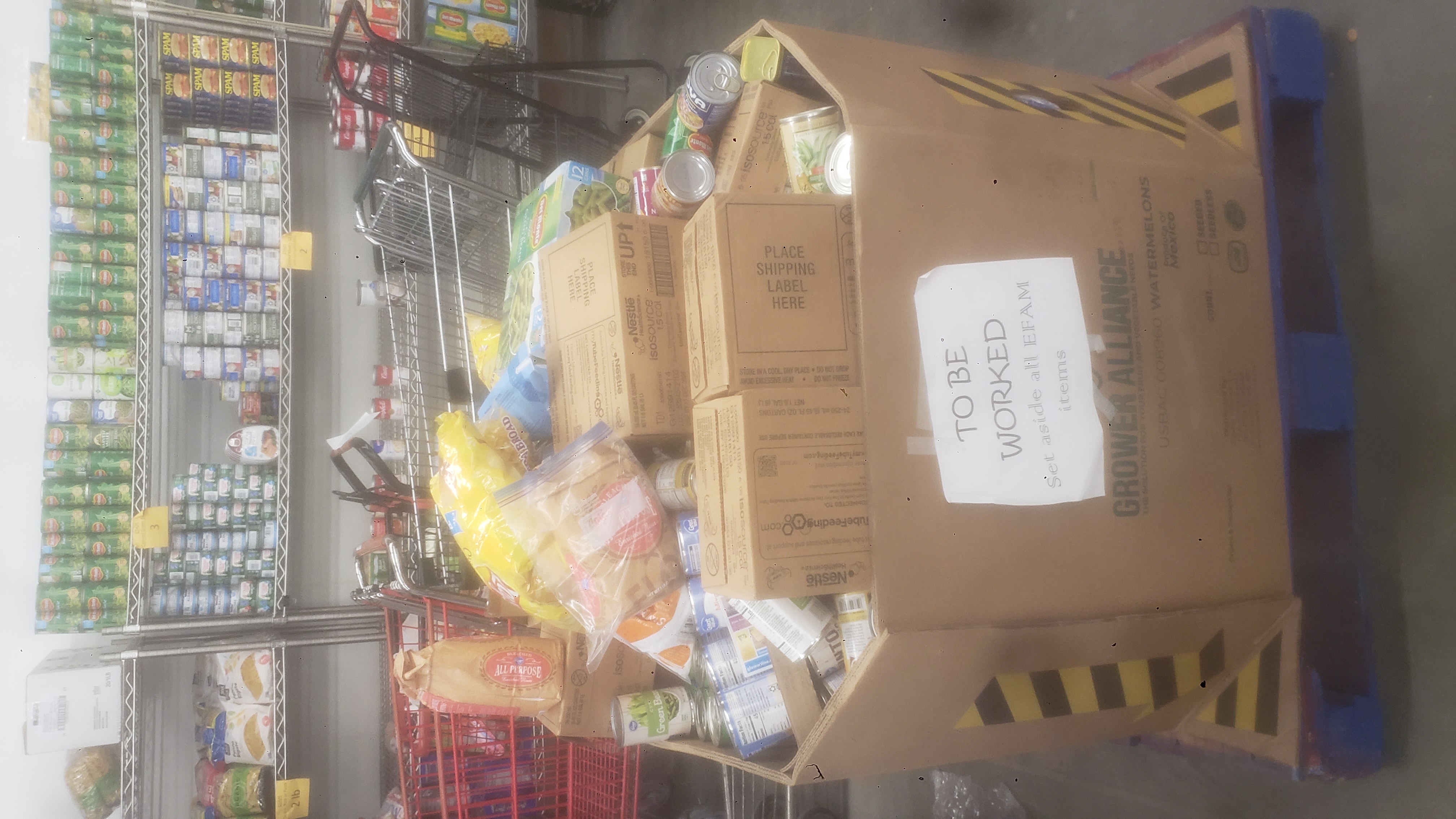 Cardboard container filled with food and a sign that says to be worked, set aside all EFAM items with shopping carts and shelves of food behind it.