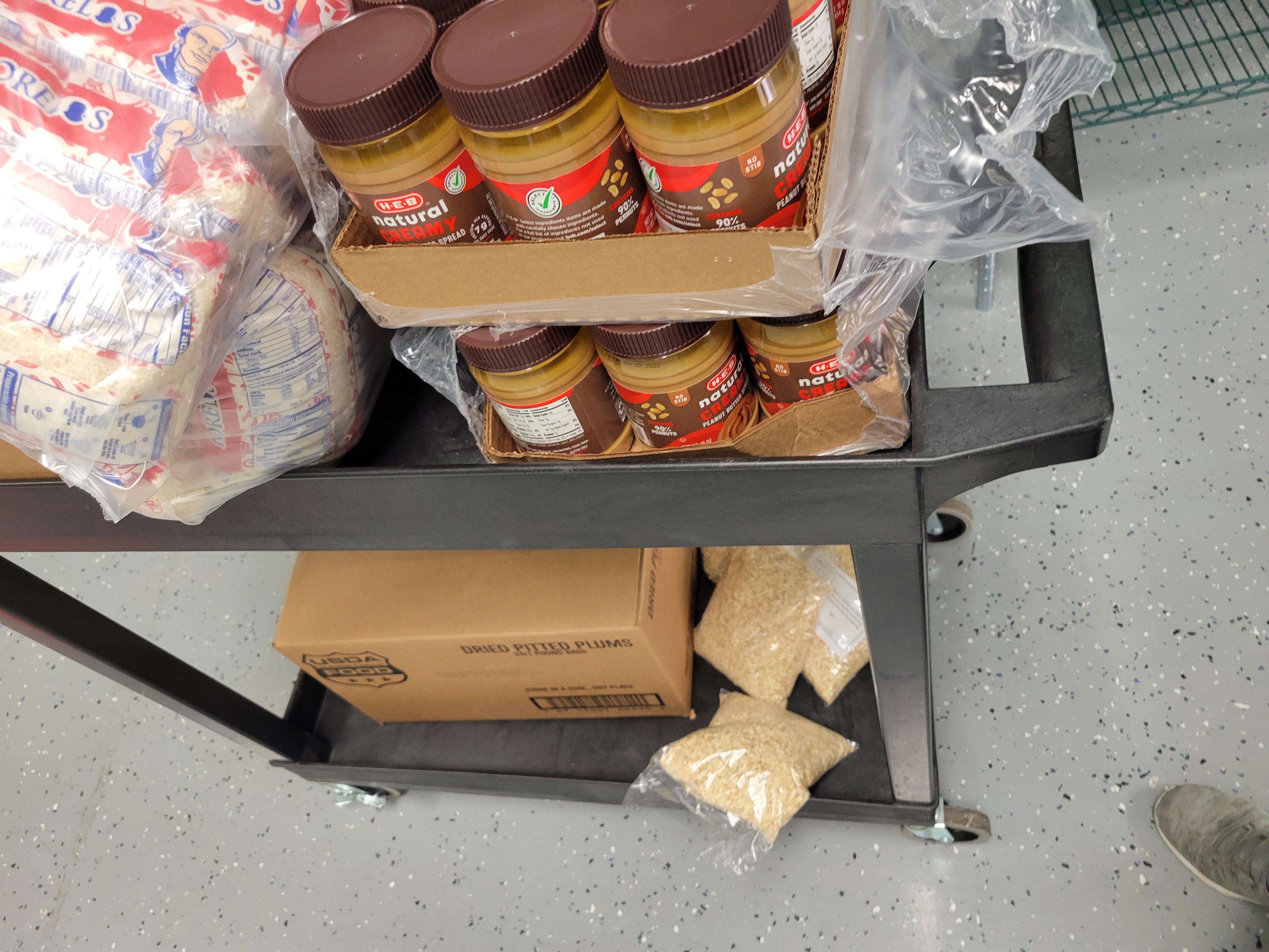 Jars of peanut butter and bags of rice on the top shelf plus bags of plums and bag of oats on bottom shelf of cart.