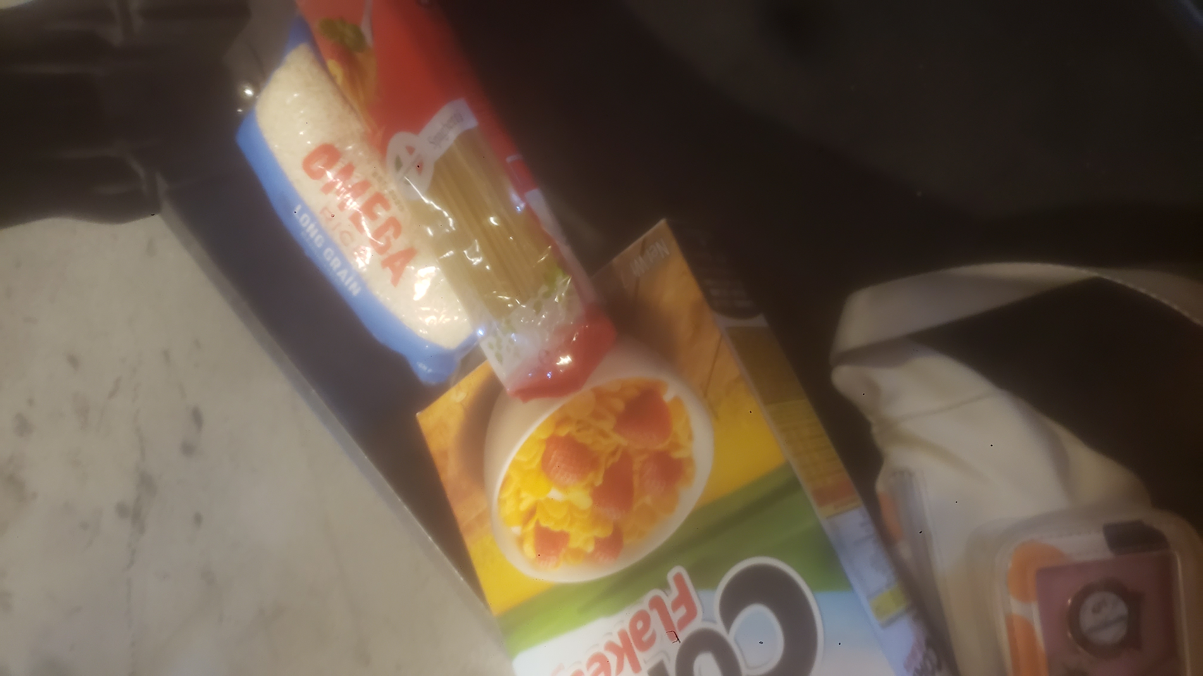 Box of corn flakes, bag of spaghetti, and bag of rice on the bottom of a shelf cart.