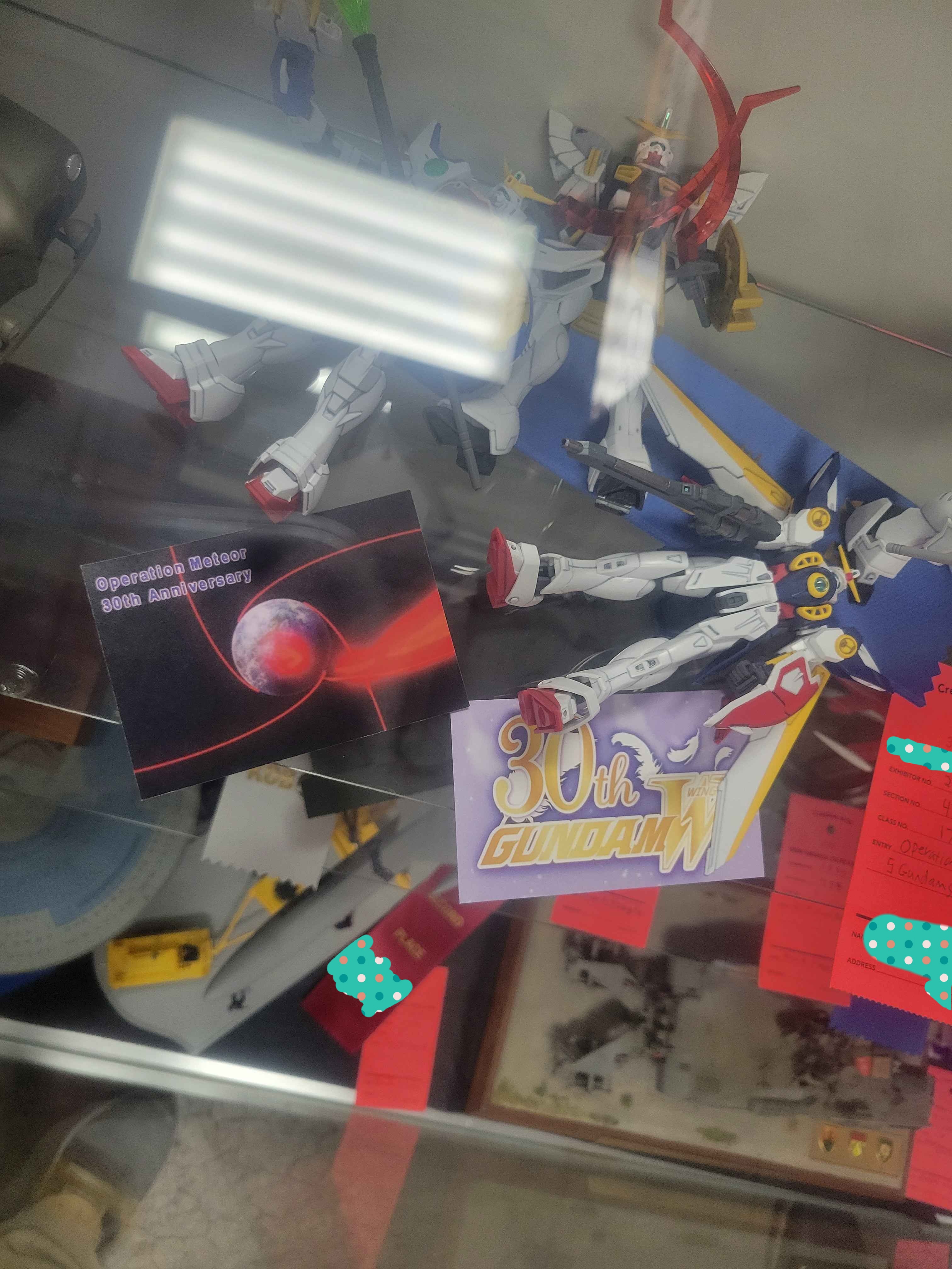 Gundam wing toys in display case with a 30th Gundam Wing anniversary card next to them.