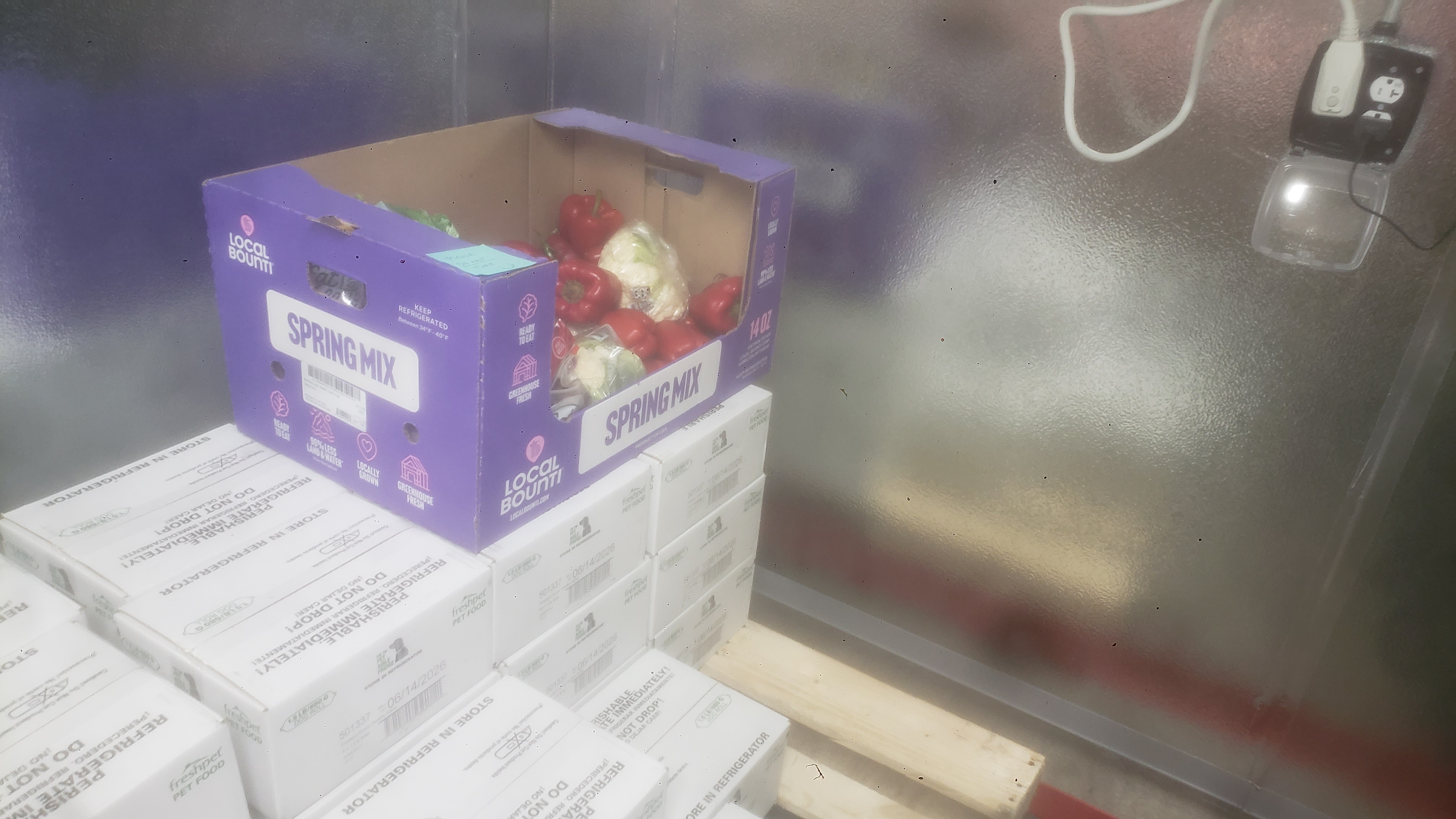 Cardboard produce crate filled with red bell peppers and packaged lettuce sitting on boxes of pet food.
