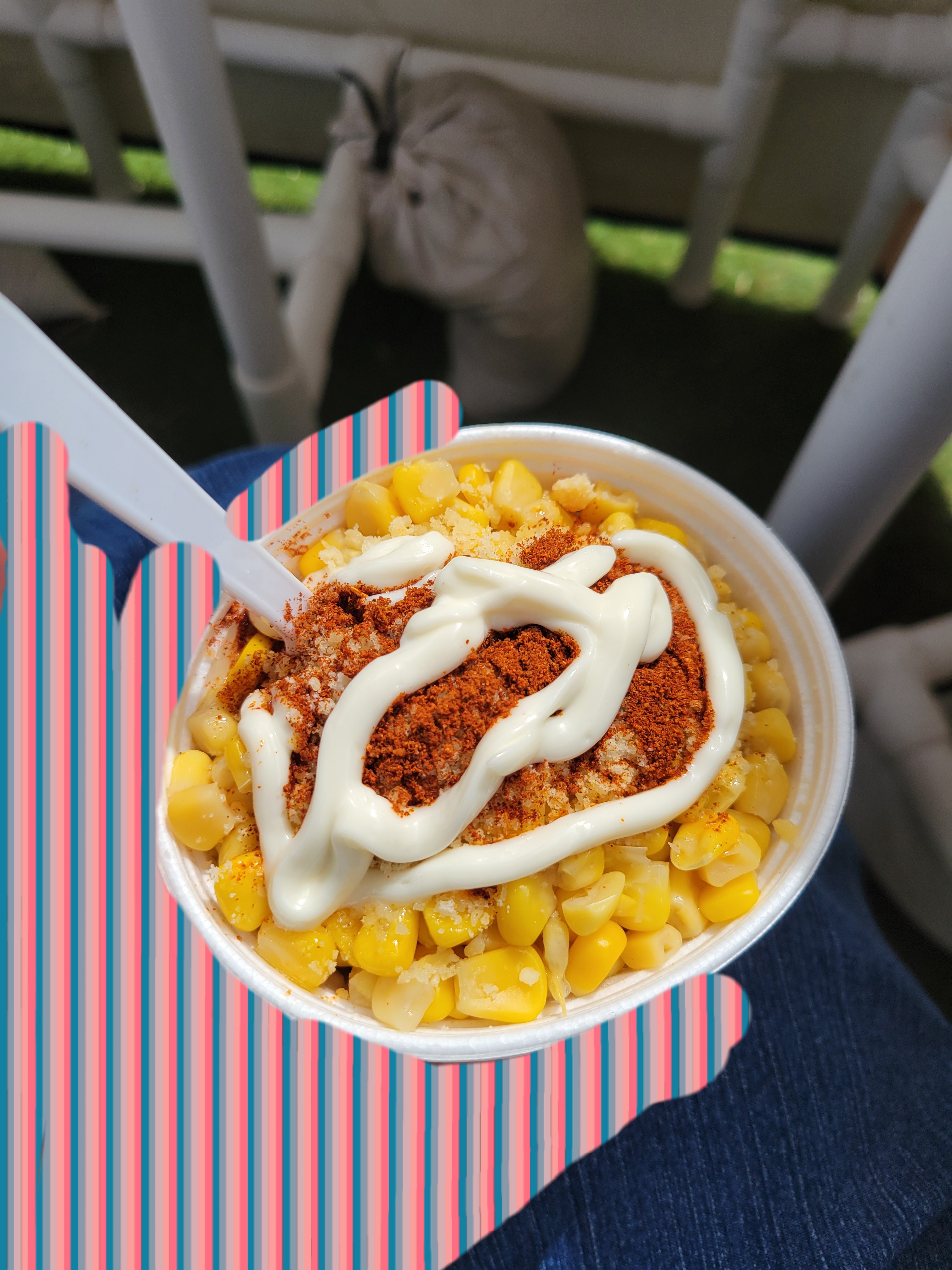 Hand holds elotes in a cup topped with mayonnaise, chili powder, and a spoon.