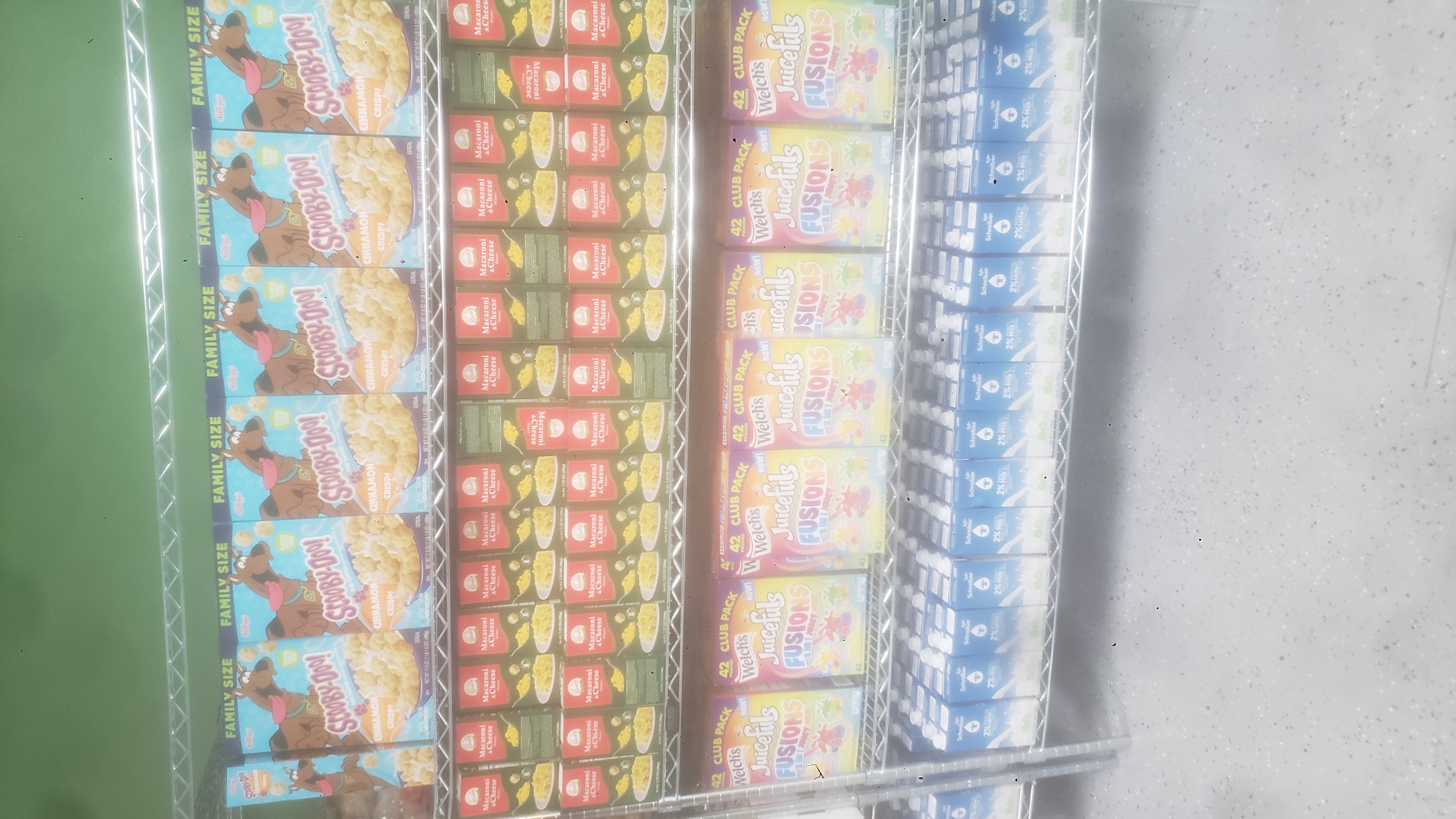 Boxes of cinnamon crisp cereal, boxes of mac n cheese, boxes of fruit snacks, and cartons of 2% reduced fat milk on a four tier shelf.