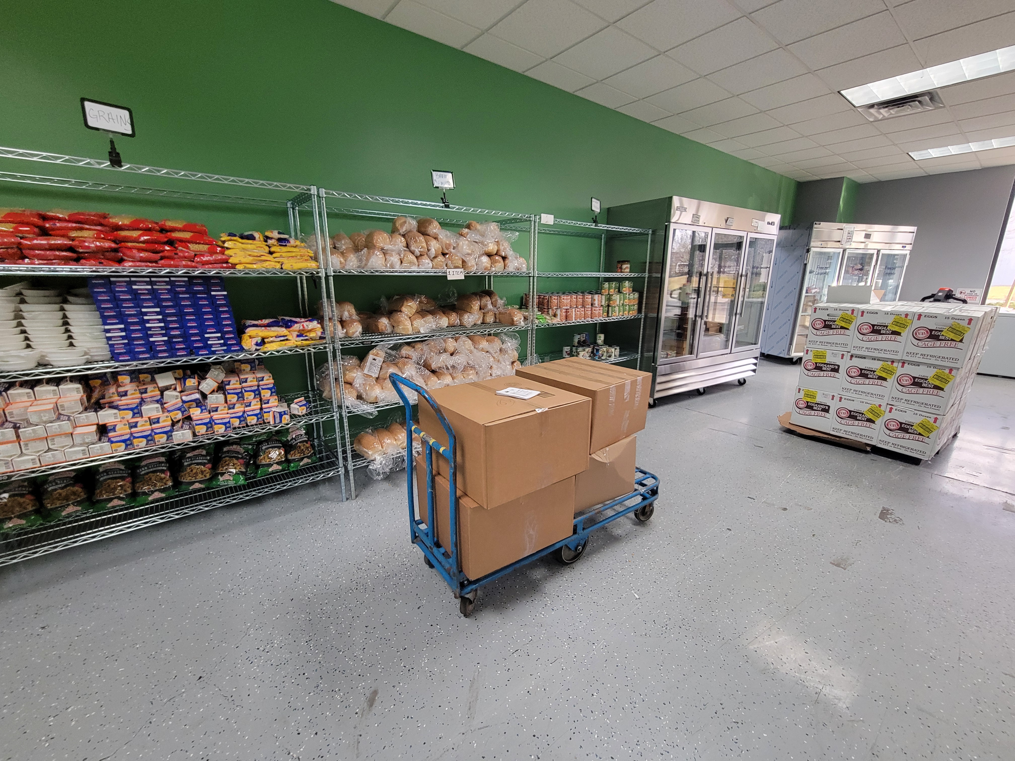 Food filled shelves with signs listing the different food groups next to fridge and boxes filled cart in front of them.