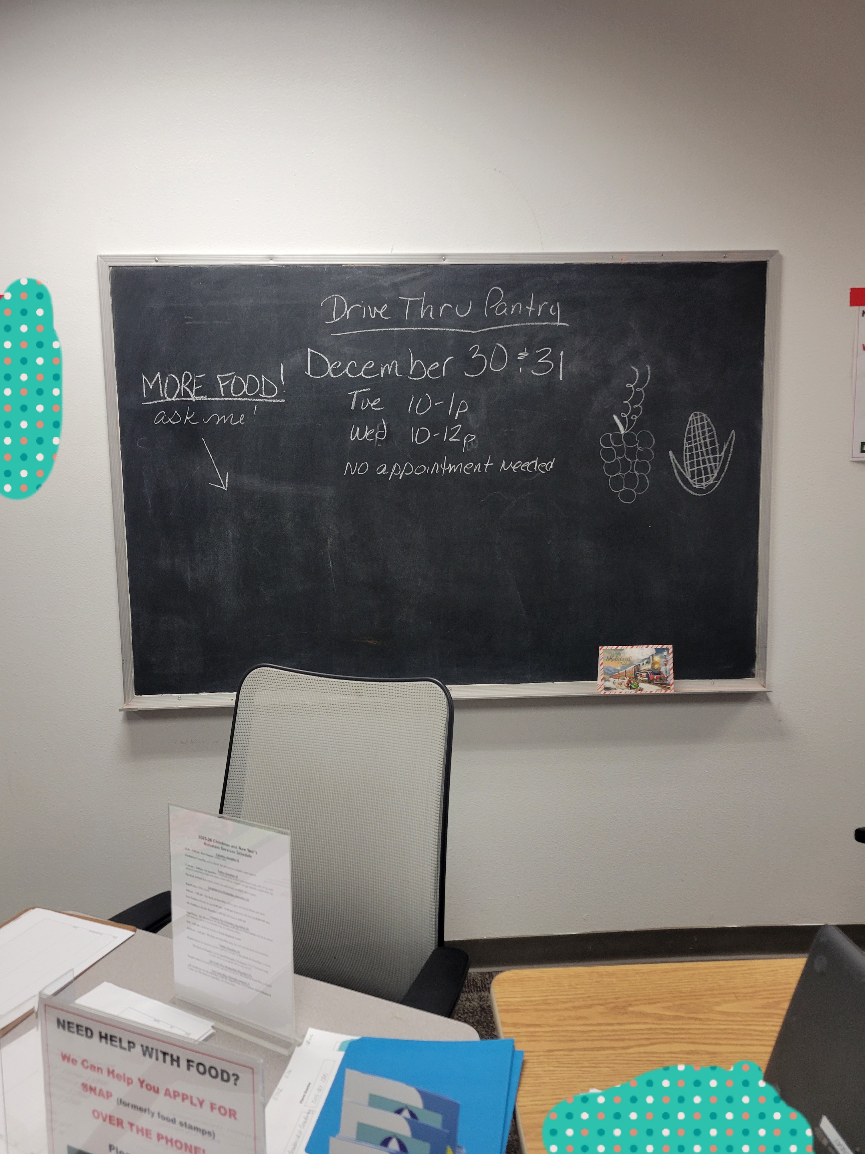 Chalk board listing the December 30 and 31 dates for the drive thru pantry with a table filled with flyers about community resources in front of it.