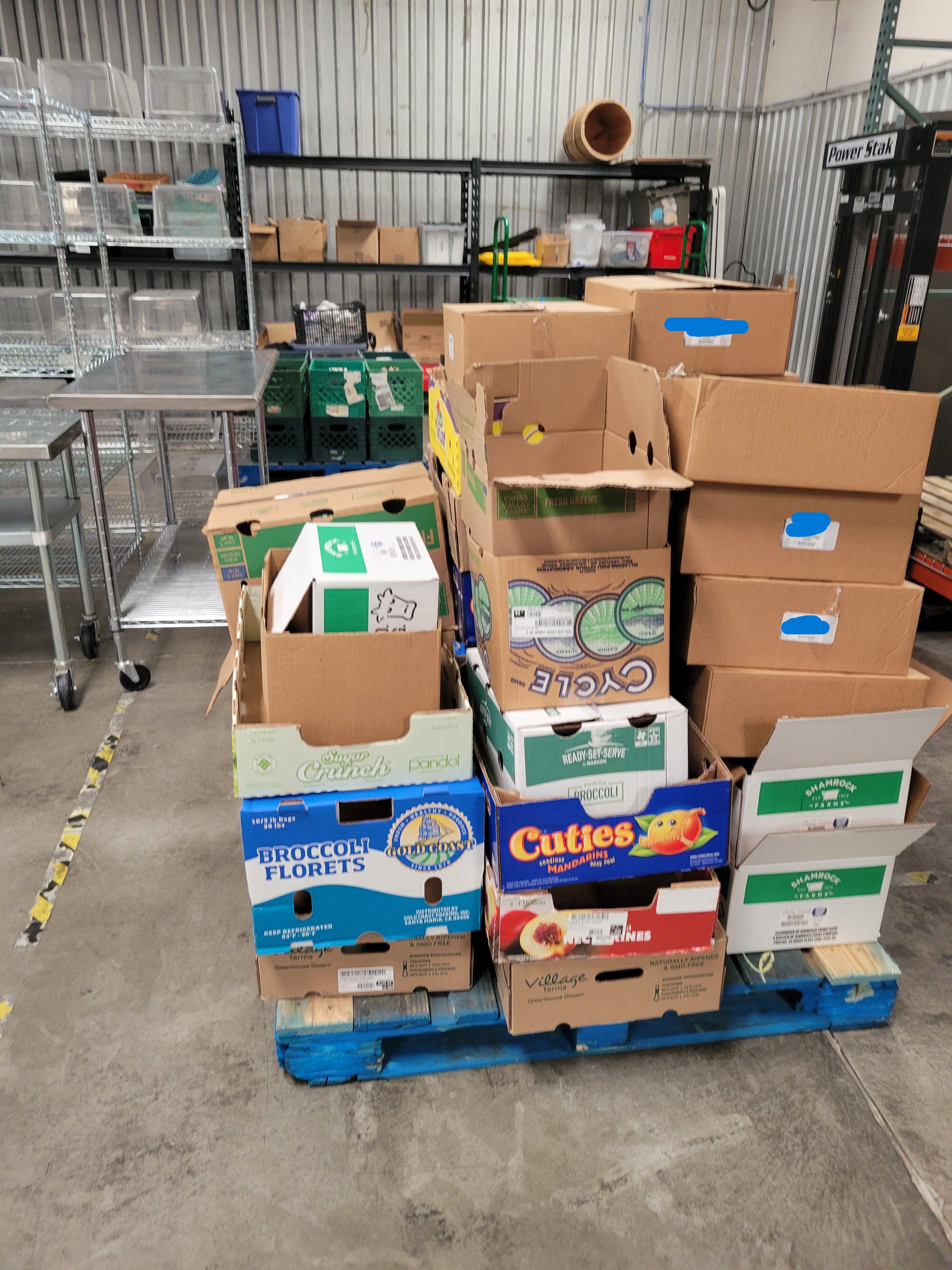 Cardboard boxes on pallet next to pallet stacker and tables plus shelves behind it.