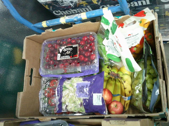 Produce box filled with container of cherries, bag of cucumbers, bag of sweet peppers, bag of green grapes, container of tomatoes, bag of salad, apples, and a bunch of bananas.