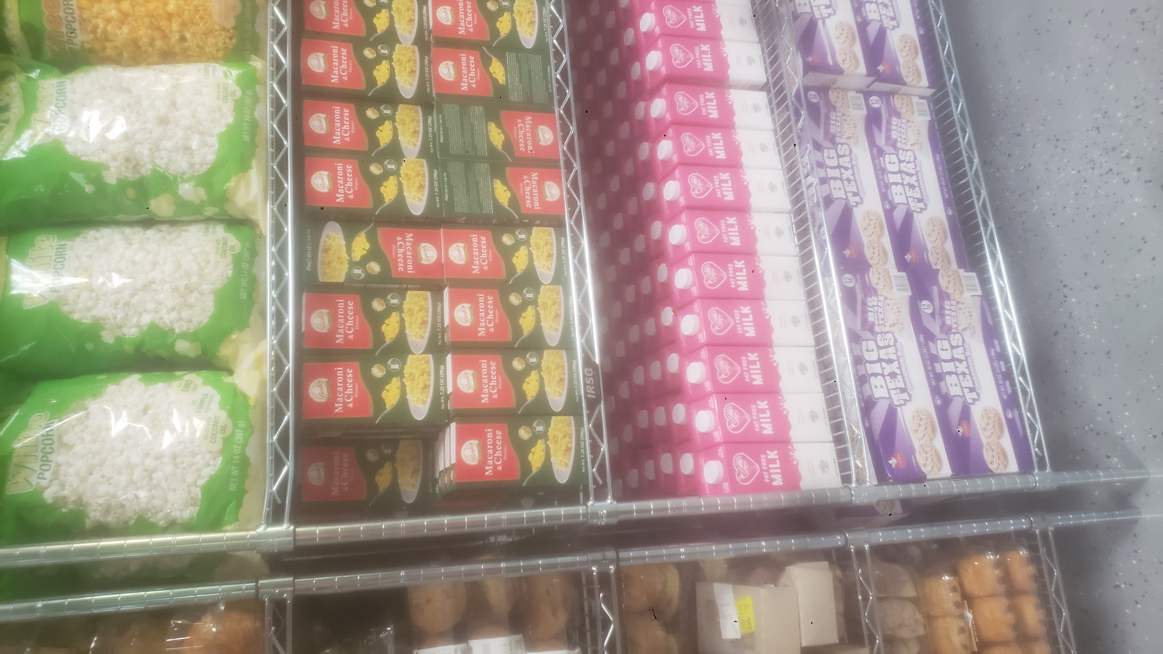 Boxes of mac n cheese, boxes of cinnamon rolls, cartons of fat-free milk on a shelf, bags of garlic parmesan chips, cheese popcorn, and white popcorn on a shelf.