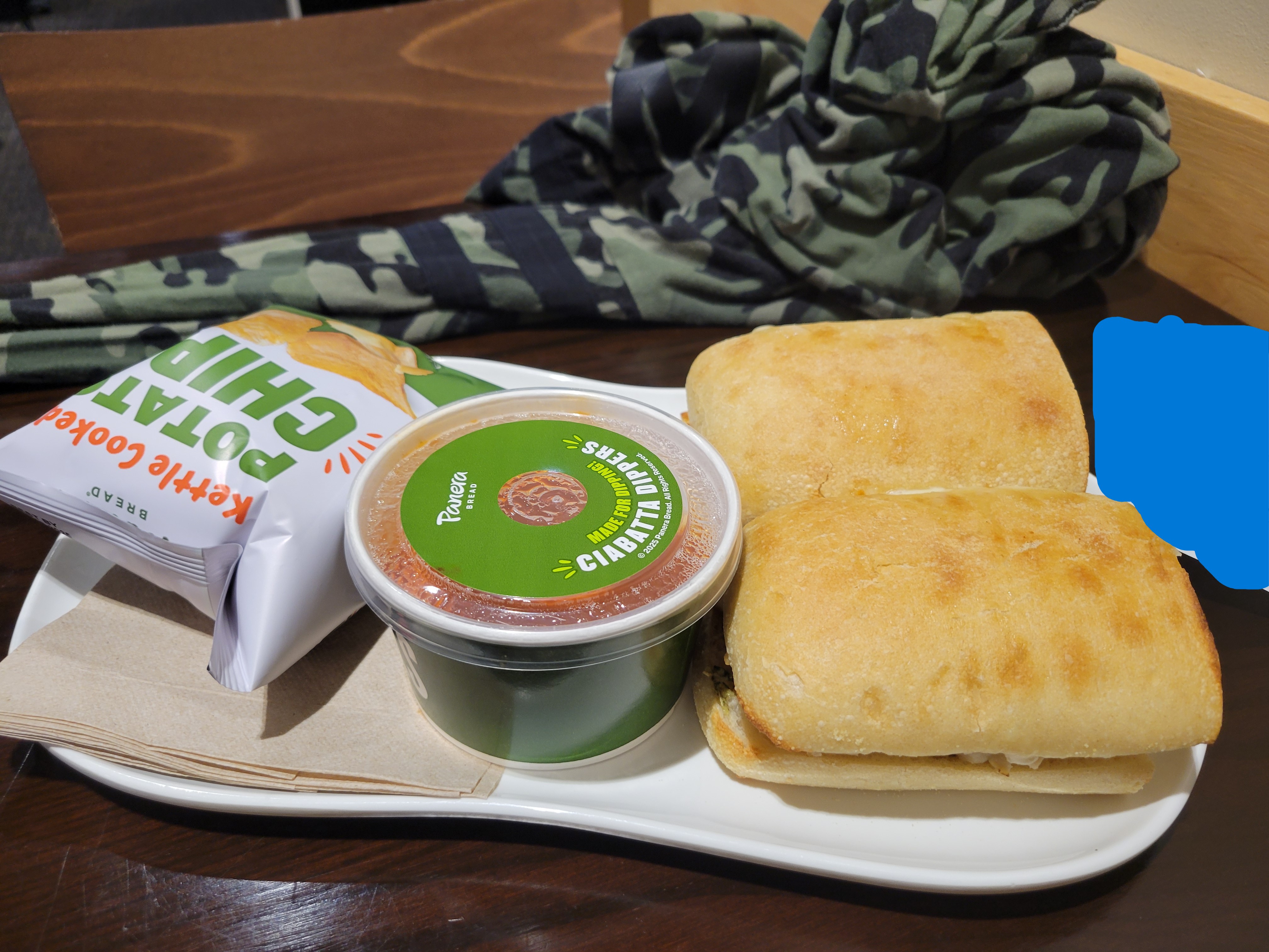 Bags of potato chips, container of soup and sandwich sits on plate.