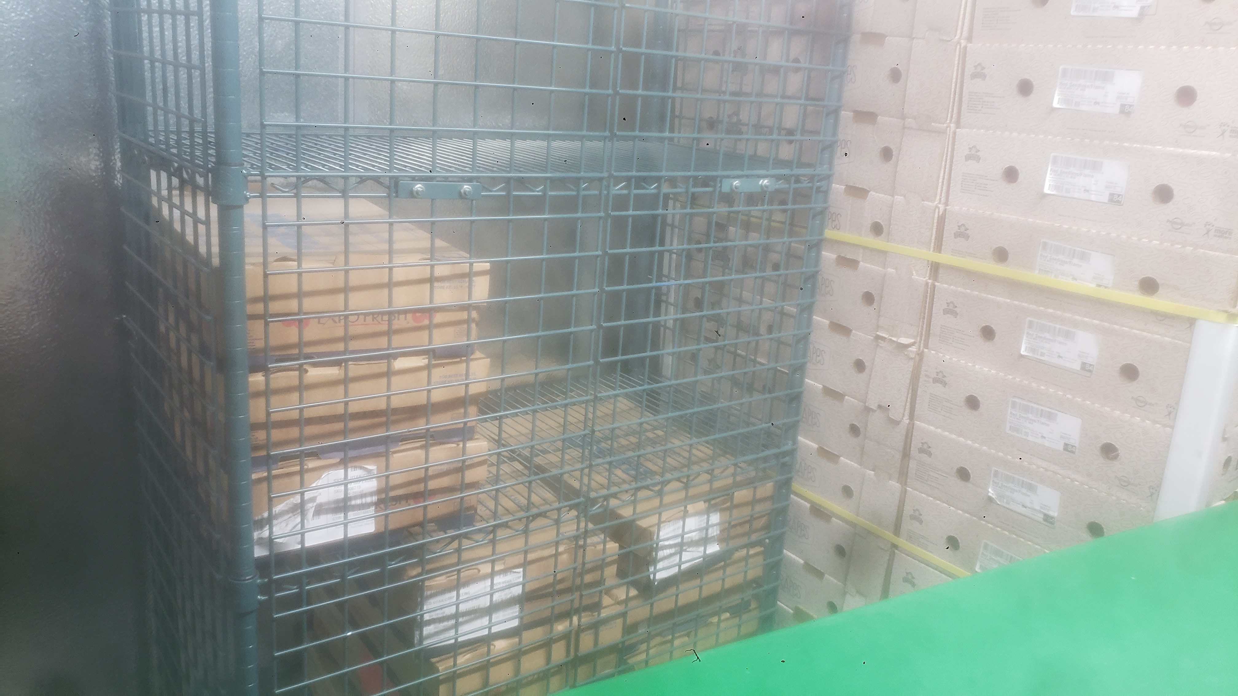 Cardboard boxes in a cage shelf next to a shelf cart and a pallet of cardboard trays.
