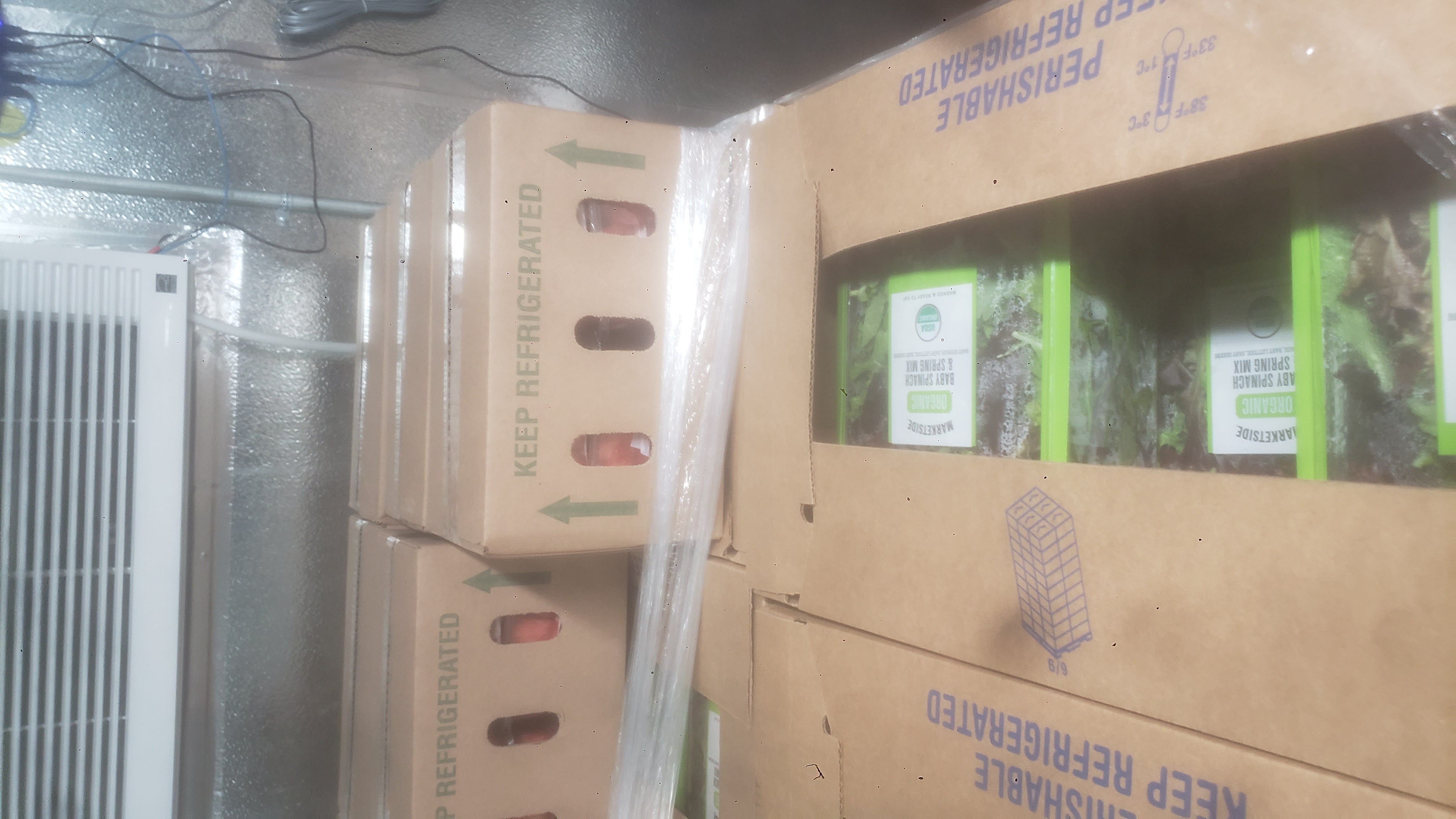 Cardboard boxes filled with containers of baby spinach and spring mix with cardboard boxes full of containers of fruits behind it.