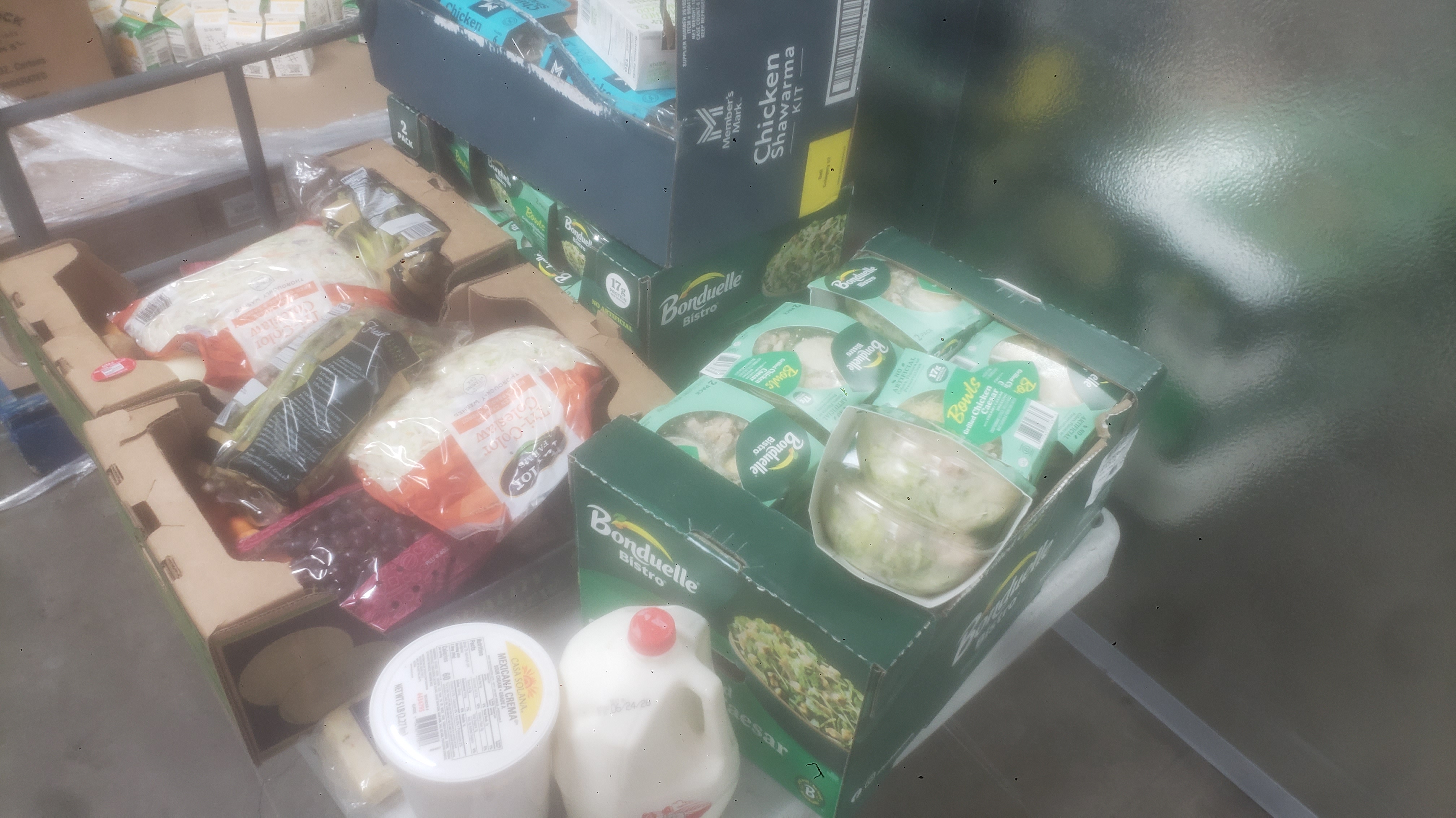 Produce trays filled with bags of produce and salads next to a gallon of milk, packet of cheese, and 5-pound container of Mexican-style sour cream.
