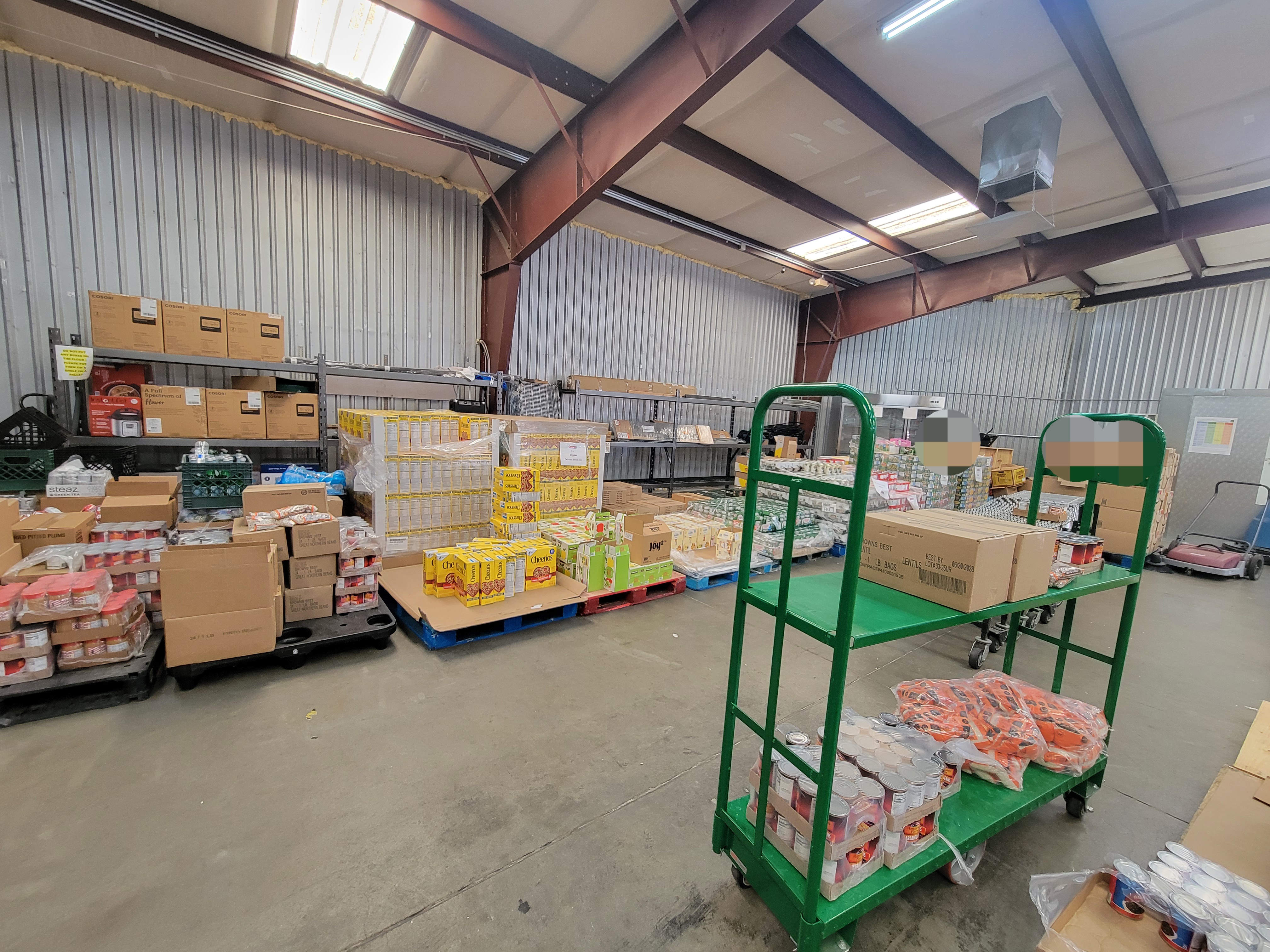Warehouse with pallets of food, shelf cart filled with food, fridges, conveyor belt, shelves, and push sweeper.