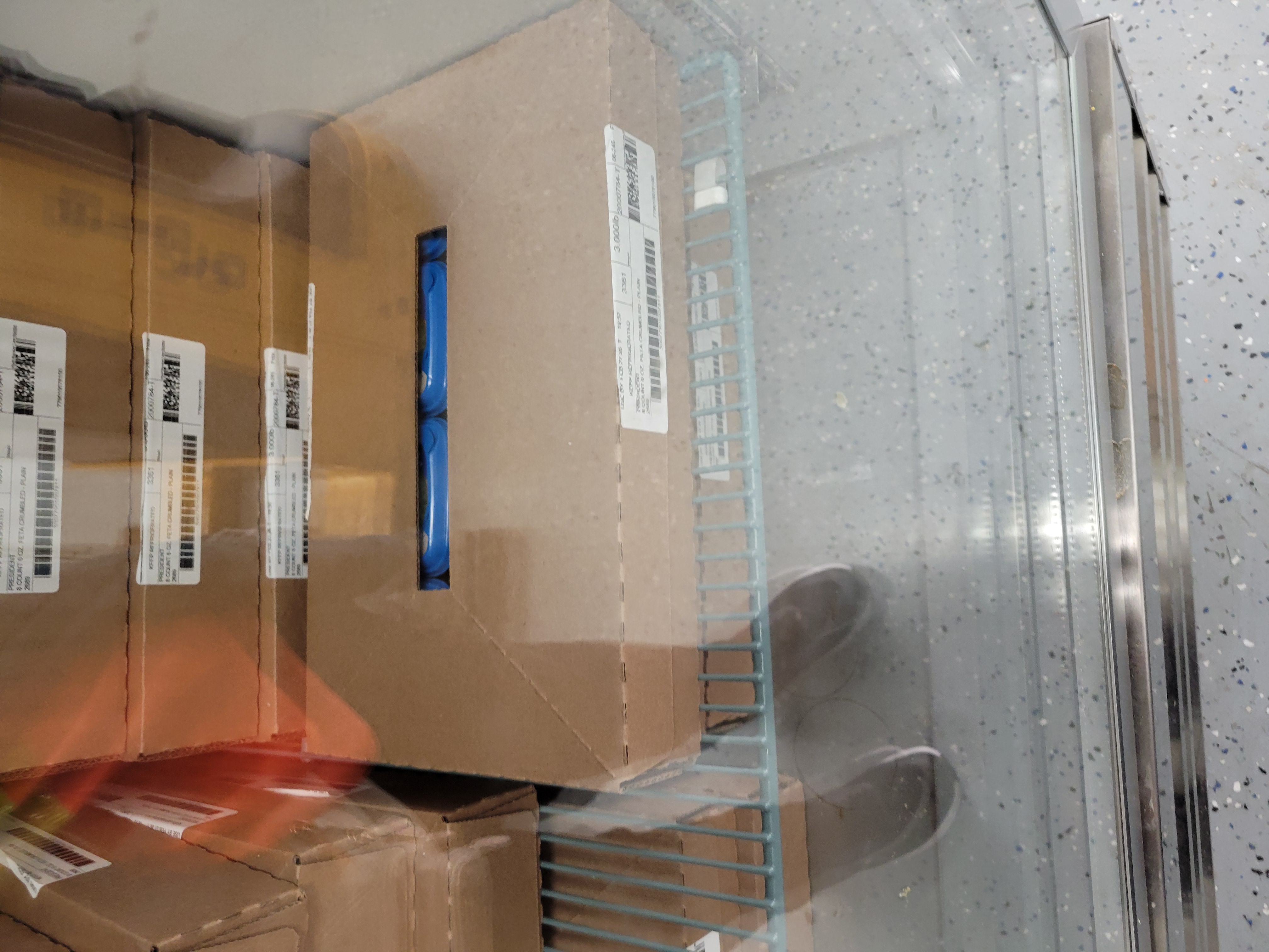 Cardboard boxes of containers of crumbled feta cheese in a fridge.