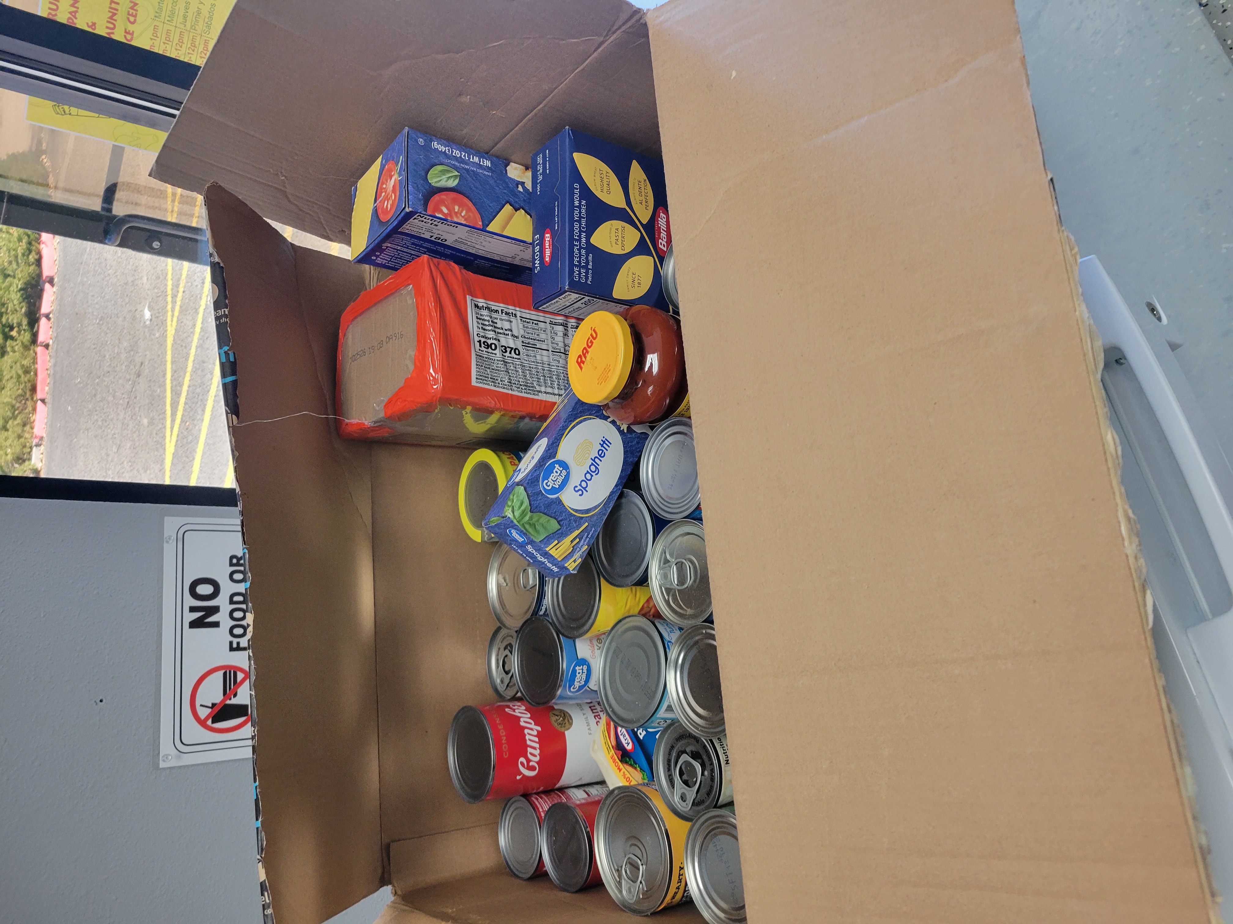 Amazon box filled with canned food, bottle of mayo, pack of ramen noodles, jar of pasta sauce, and different noodles.