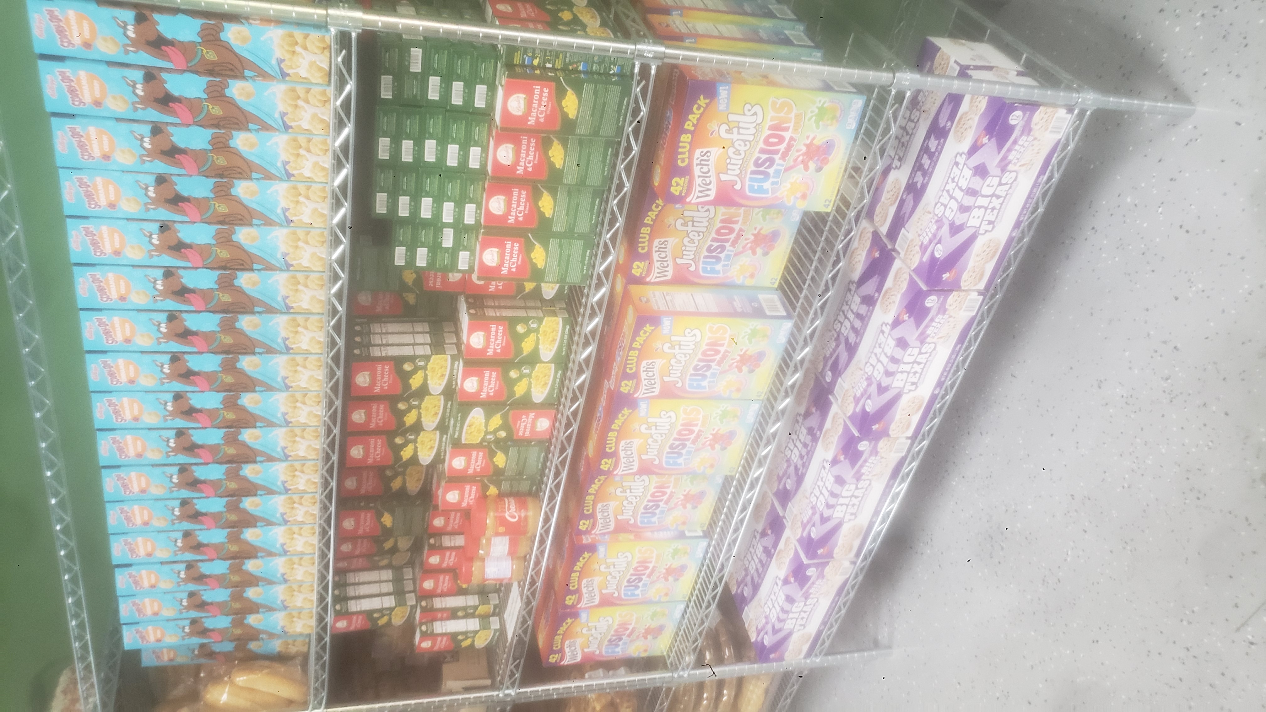 Boxes of cinnamon crisp cereals, boxes of mac n cheese, jars of peanut butter, boxes of fruit snacks, and boxes of cinnamon rolls on a shelf.