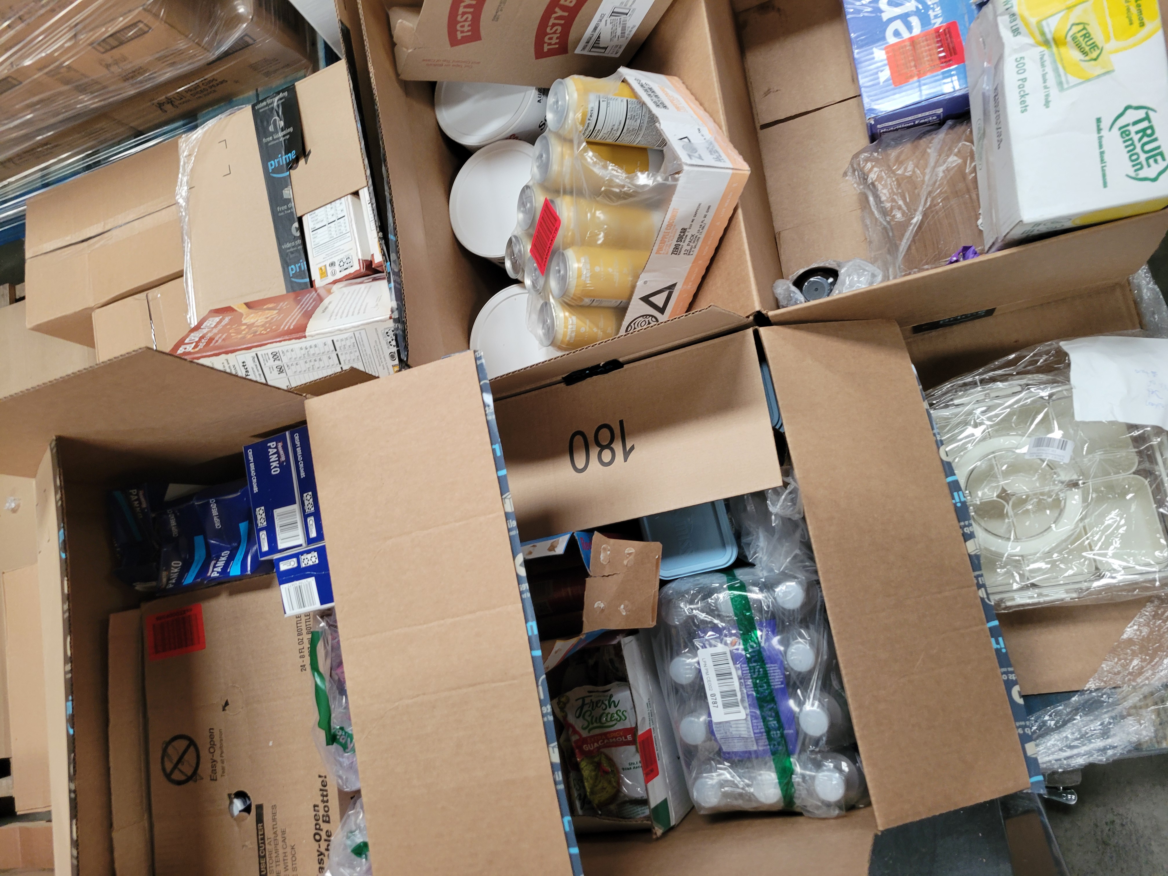 Two boxes, box of oil and one bag of chips in fruit box on pallet next to shopping cart filled with food.