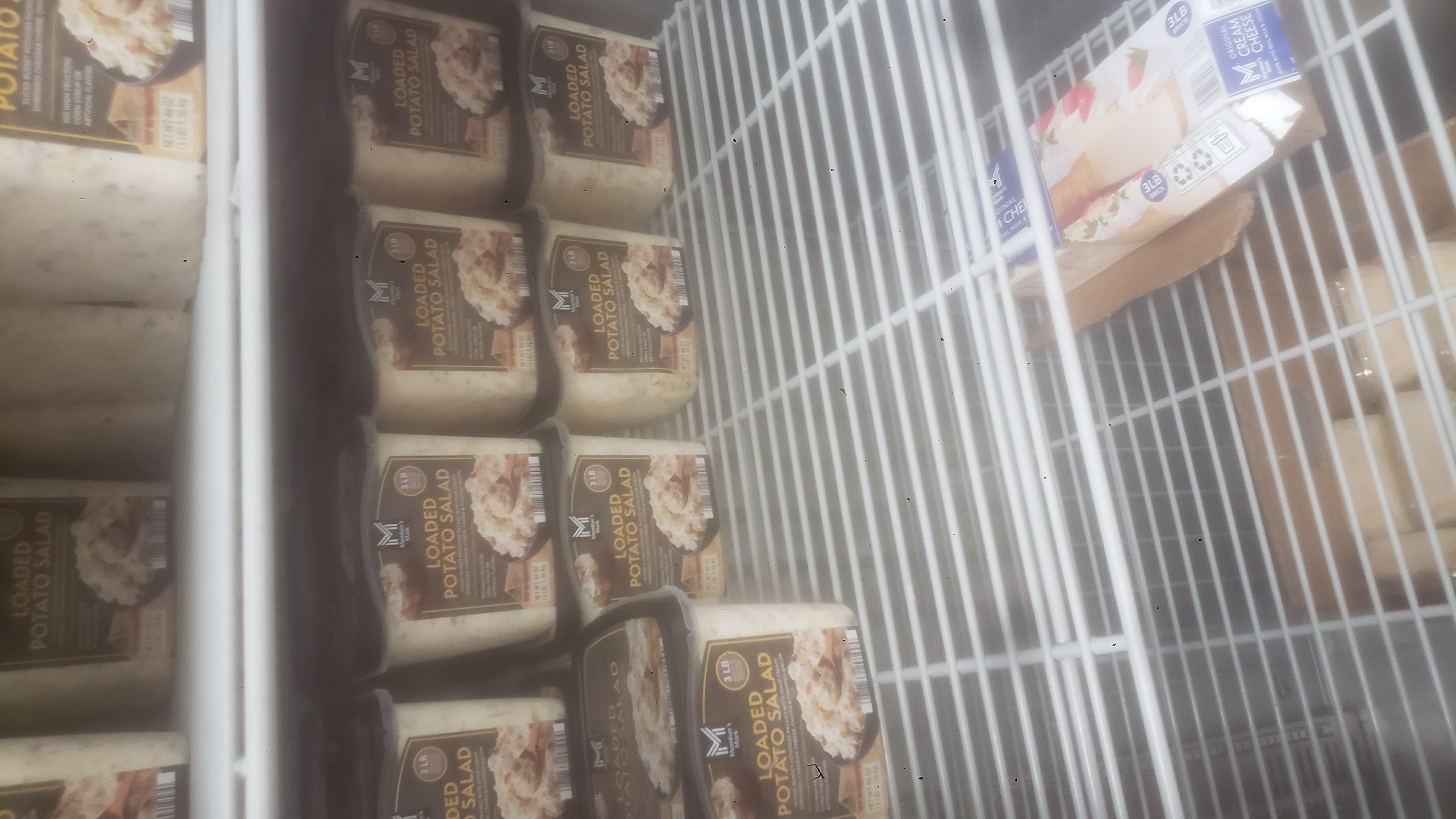 Containers of loaded potato salad and cardboard box of cream cheese on shelves of a fridge.