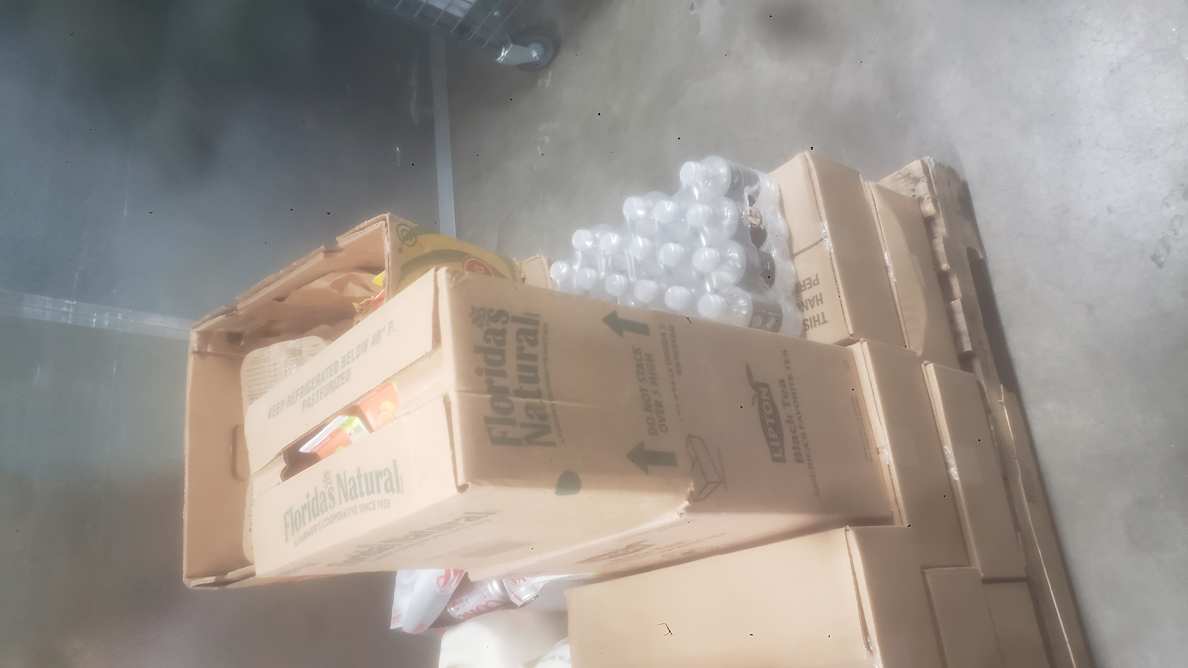 Case of water bottles, box of black tea, case of diet coke, box of orange juice, and banana box full of miscellaneous things on top of cardboard boxes.