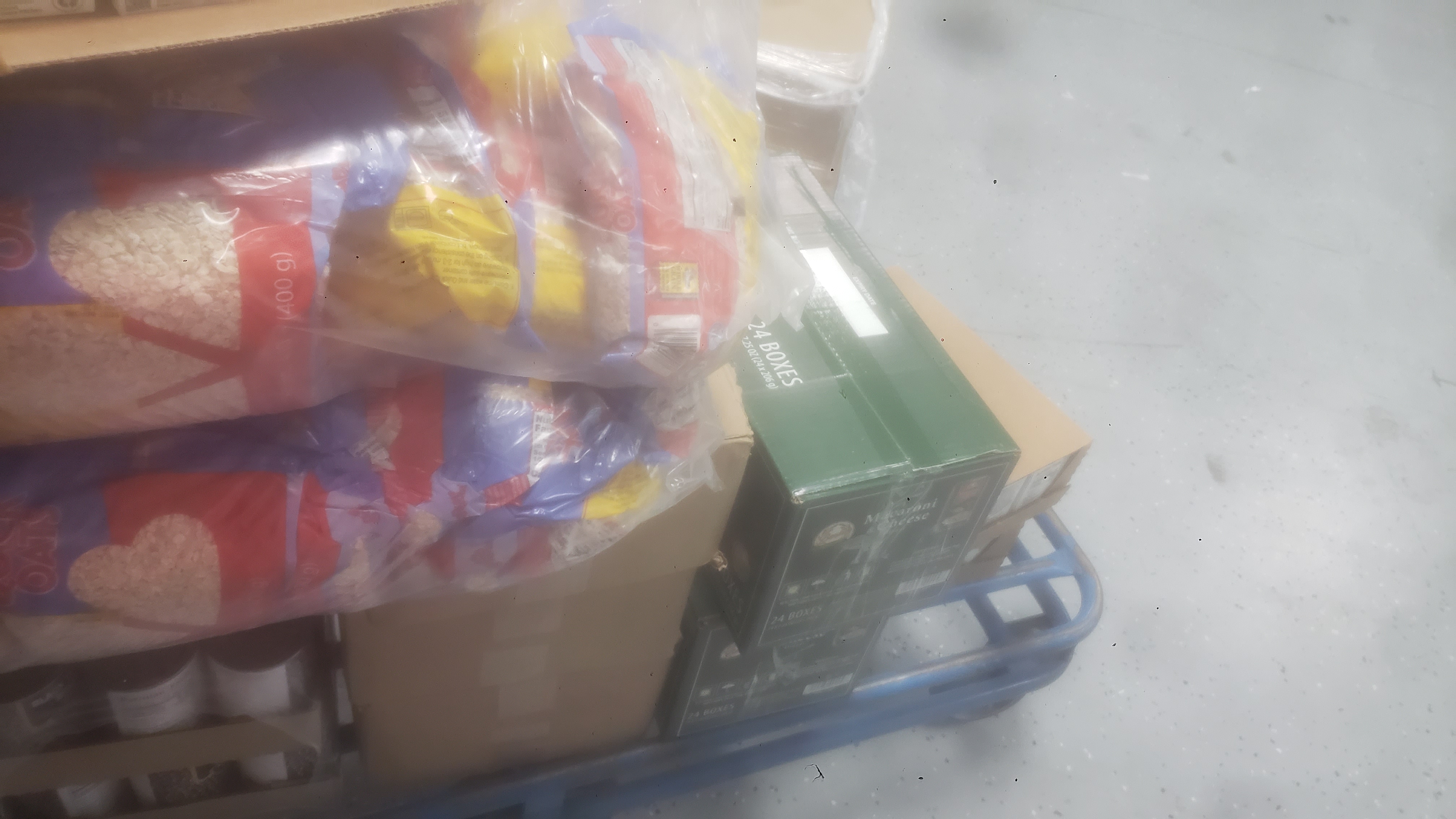 Box of shelf-stable meals on top of bags on oats. Under that is cans of tomatoes sauce, applesauce, and black turtle black beans which is next to boxes of mac n cheese and shelf-stable meals on a shelf cart.