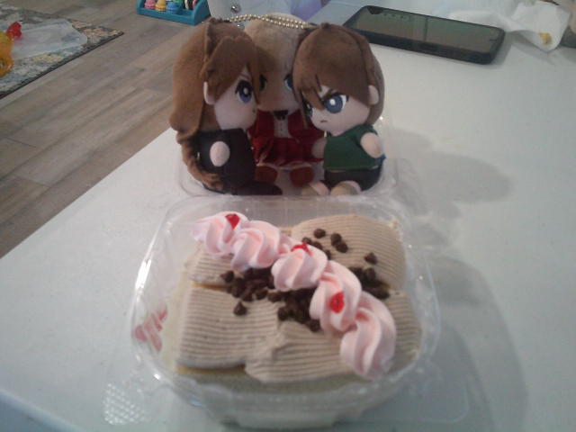Gundam wing Relena, Heero, and Duo plush on the top of a to go container of a tres leches cake .
