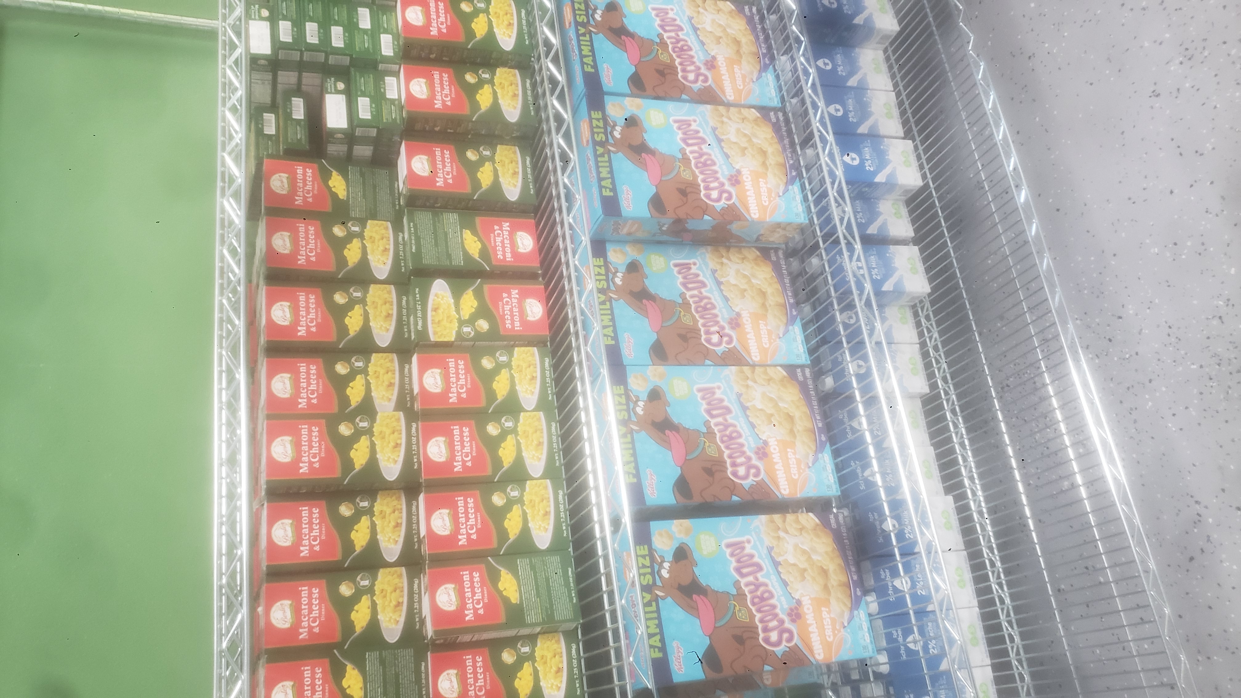 Boxes of mac n cheese, boxes of cinnamon crisp cereals, and 2% reduced milk cartons on racks of a shelf.
