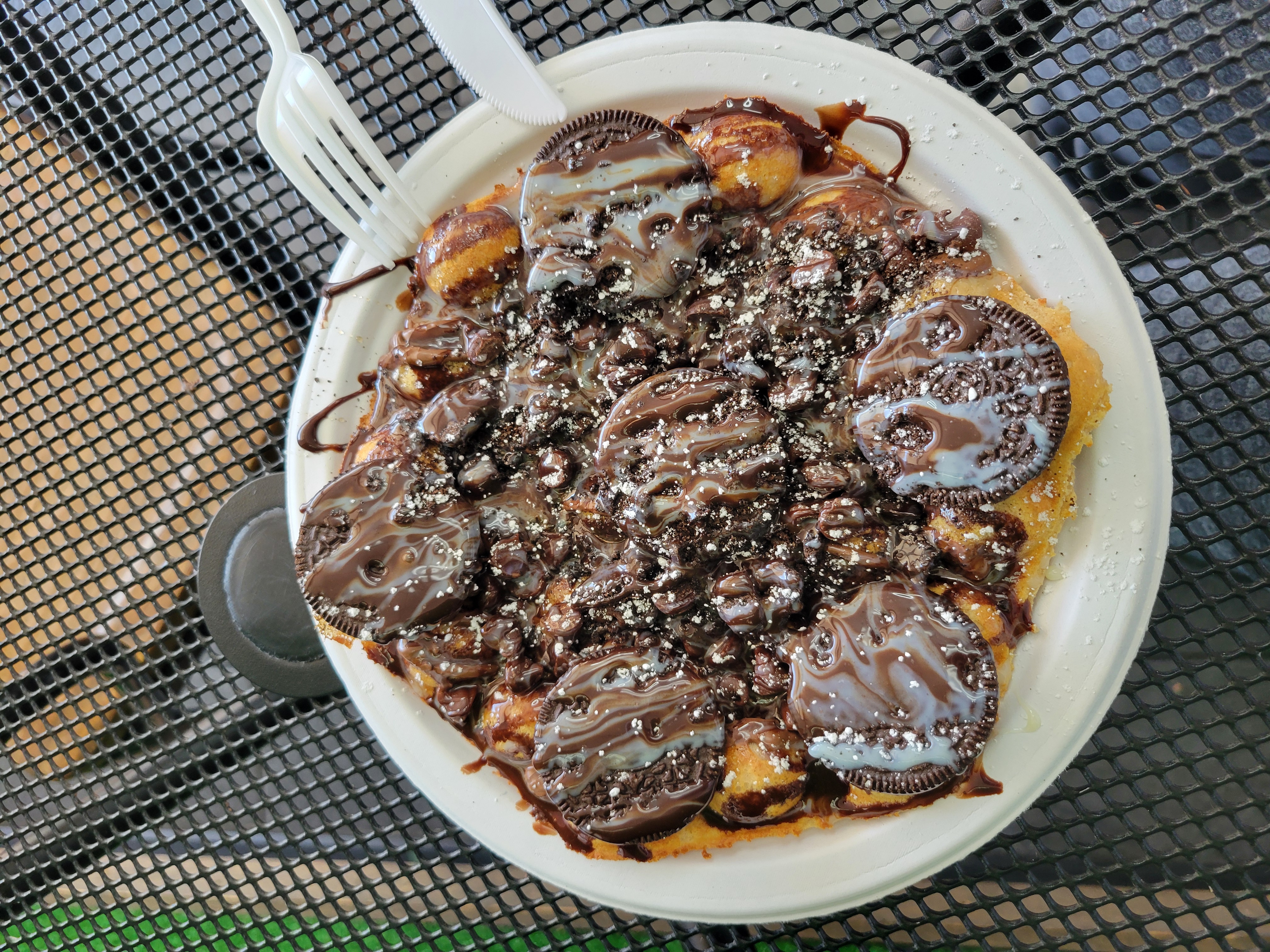 Chocolate sauce and oreo bubble waffle on a paper plate with a plastic knife and fork on the edge of the plate.
