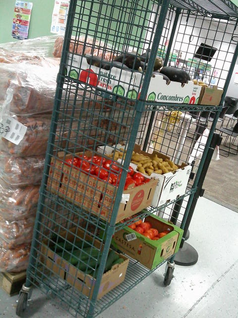 Cage shelf filled with produce boxes of tomatoes, cucumbers, and potatoes next to a wrapped pallet of carrots.