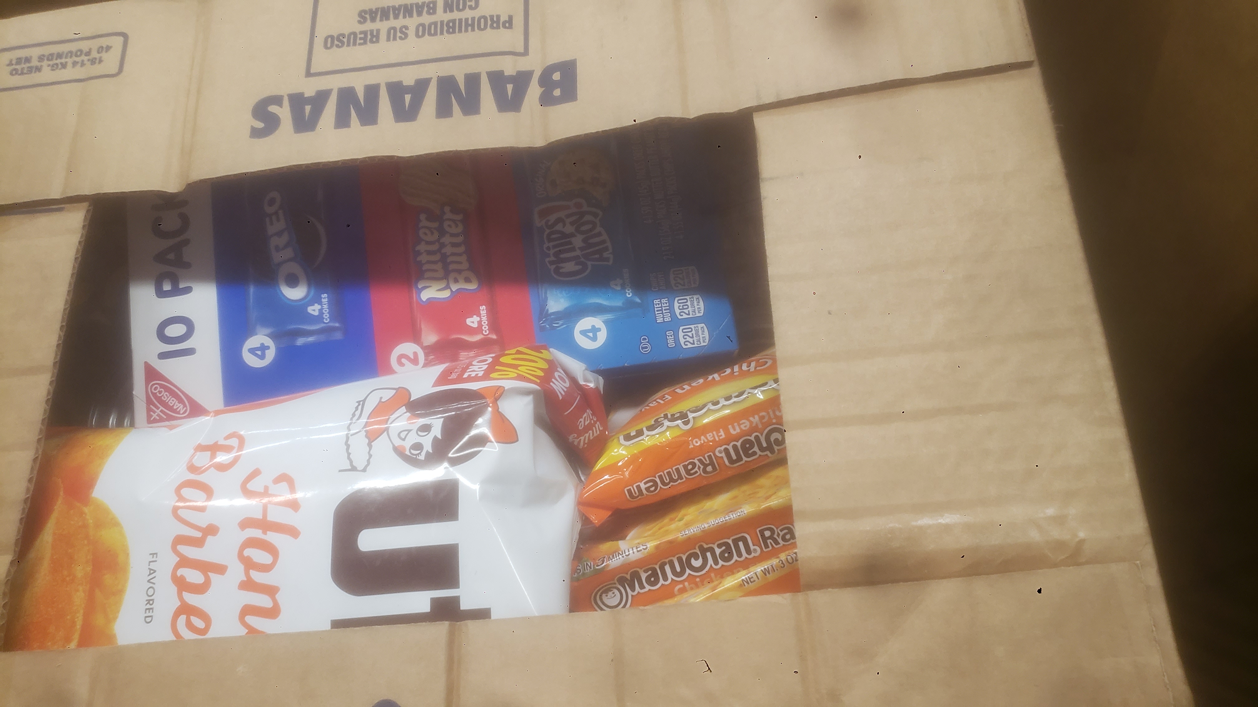 Banana box of emergency food with two packs of ramen noodles, bag of honey barbecue chips and box of different cookies showing on top.